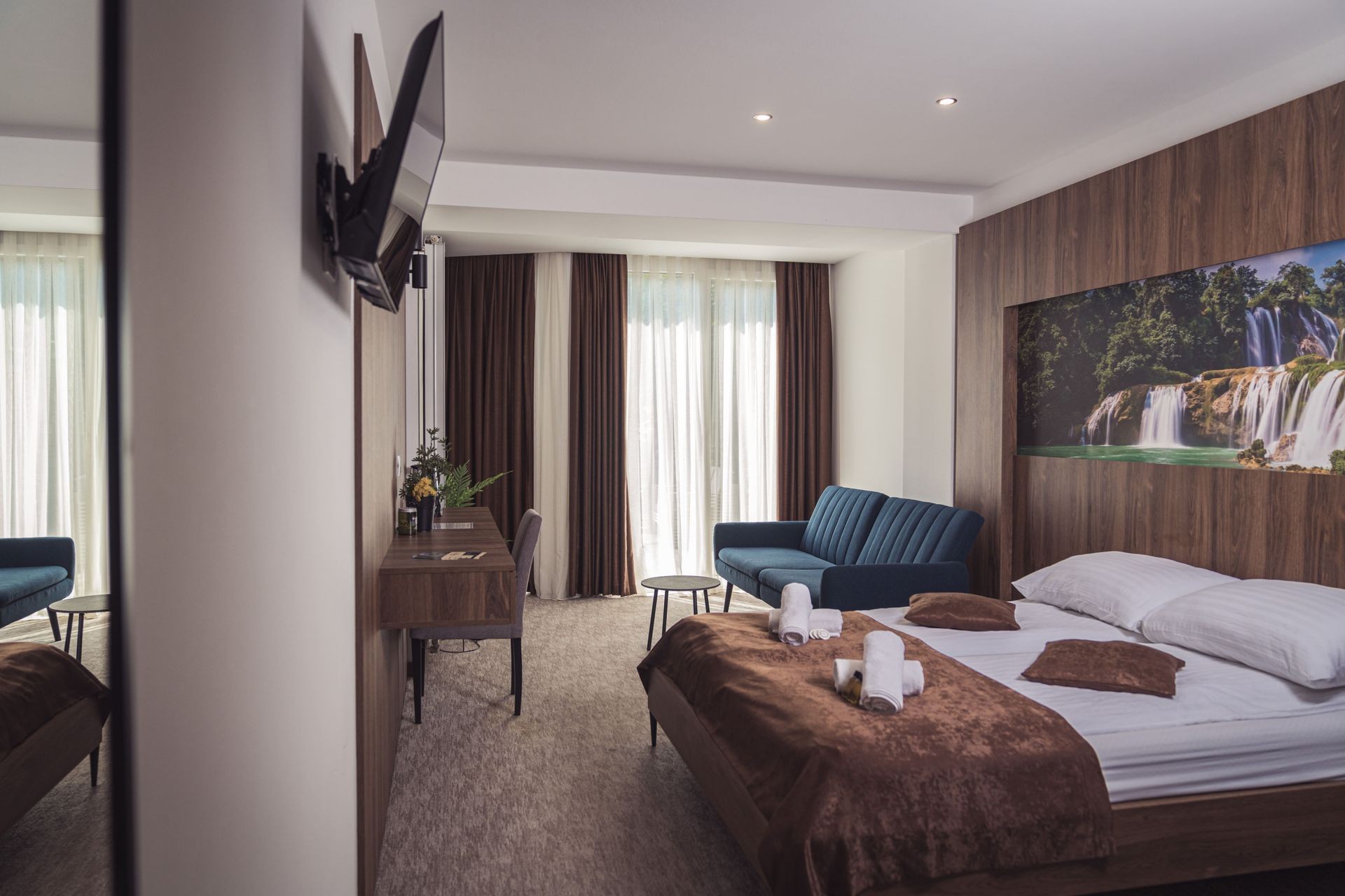 "Spacious hotel room in Hotel Tazz in Bihać with double bed, sofa, desk, and waterfall-themed decor"